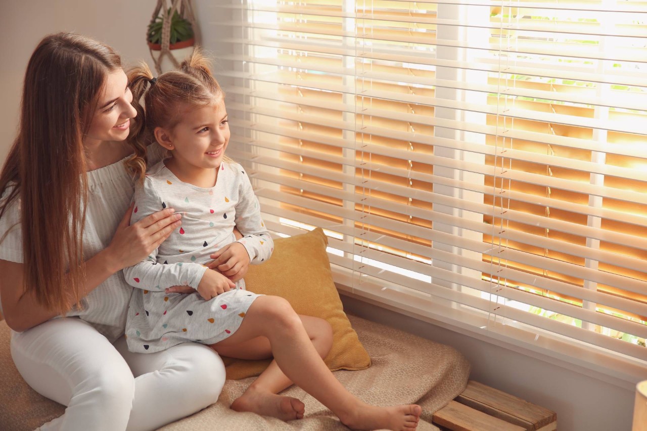 A mother sitting with her daughter looking out their blinds with no exposed cords or wands at Dale’s AAA Blind Factory near Naples, Florida